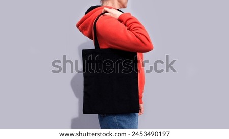 Woman in red hoodie holding tote canvas blank eco bag on street grey minimal wall background. Female consumer hold black textile shopper. template or place for your design, logo, text