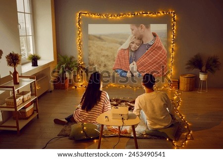 Young couple enjoying Valentine's Day, having romantic date at home, having good time together, watching movie on modern projector in cozy living room interior with beautiful fairy lights. Back view
