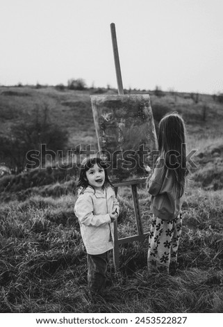 Black and white photo of two young children painting on an easel outdoors in a grassy field, one child facing the camera with a surprised expression.