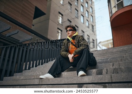 In heart of city's rhythm. Cool attitude young man sitting with thoughtful expression on stairs against skyline, urban background. Concept of street fashion and urban style, modern lifestyle, gen Z