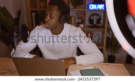 Adult african american man podcasting at night in his home office, using a microphone and laptop.