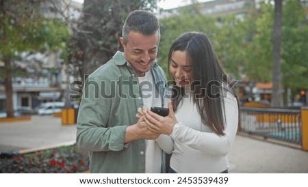 A cheerful couple shares a moment looking at a smartphone together in a city park surrounded by greenery. Royalty-Free Stock Photo #2453539439