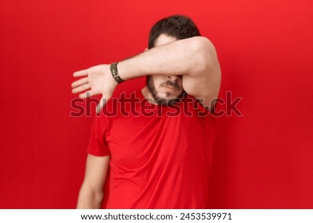 Young hispanic man wearing casual red t shirt covering eyes with arm, looking serious and sad. sightless, hiding and rejection concept  Royalty-Free Stock Photo #2453539971