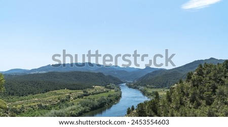 Serene river winding through a verdant valley flanked by rolling hills and lush forests under a clear blue sky, showcasing nature's tranquility