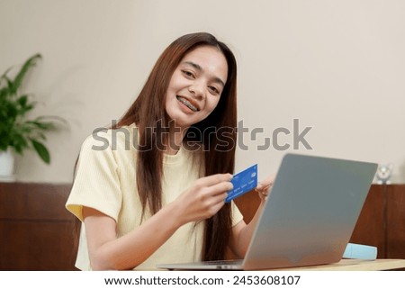Radiant asian lady with braces presents credit card, engaging in e-commerce with a portable computer at home. Delighted consumer 
