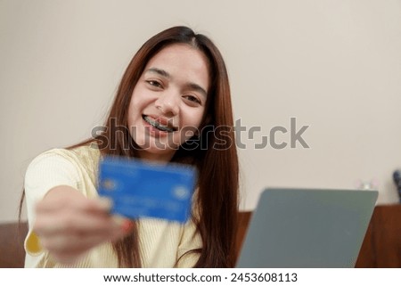 Radiant asian woman with braces presents credit card, engaging in e-commerce with a portable computer at home. Delighted consumer showcases a blue card, about to shop online using her laptop