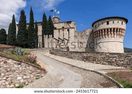 Towering cypress trees flank the drawbridge leading into the historical Brescia Castle, with the Mirabella tower adorned by the Italian tricolor Royalty-Free Stock Photo #2453617721