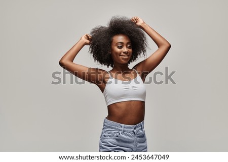 A stylish young African American woman with curly afro hair poses confidently for the camera in a studio setting.