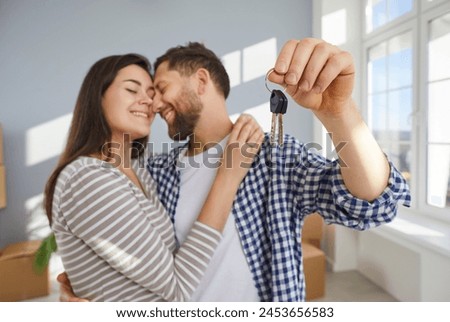 Happy young couple holding a key in hands standing in the living room at home hugging enjoying real estate or car purchase, smiling and celebrating moving day. Relocating, mortgage concept. Royalty-Free Stock Photo #2453656583