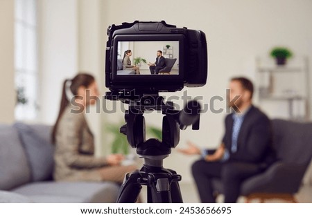 Camera on tripod foreground, businessman giving interview to journalist in office, female reporter interviewing guest speaker for podcast, radio, business talk, media show, broadcasting success story