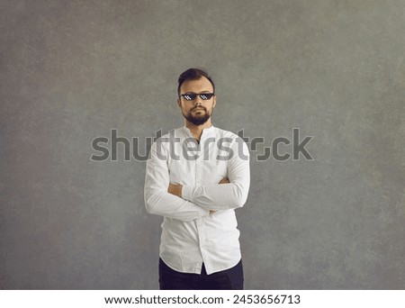 Studio portrait of guy in funny sunglasses. Serious young man in white shirt and thug life meme glasses standing arms crossed isolated on grey background