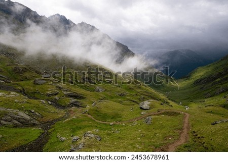 Looking in to the green valley after the storm with winding path and low hanging clouds, Eisseehütte, Hohe Tauern National Park, Osttirol, Austria Royalty-Free Stock Photo #2453678961