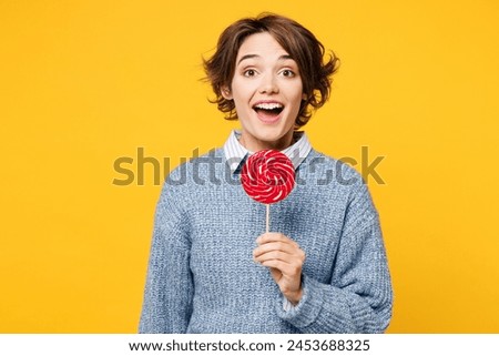 Young amazed happy fun cool woman she wears grey knitted sweater shirt casual clothes look camera hold in hand eat pink lollipop isolated on plain yellow background studio portrait. Lifestyle concept
