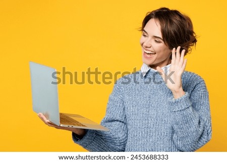 Young smiling happy IT woman she wears grey knitted sweater shirt casual clothes hold use work on laptop pc computer waving hand isolated on plain yellow background studio portrait. Lifestyle concept