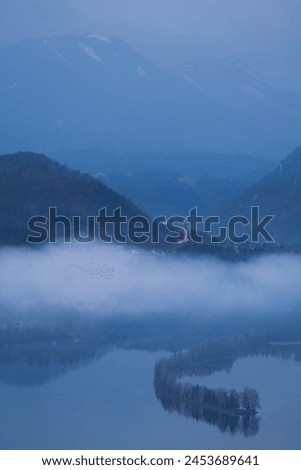 A small church lights up in a minimal scene with fog covering the village, still waters of the river Drau and towering Karawanken mountains in the distance, Carinthia, Austria Royalty-Free Stock Photo #2453689641