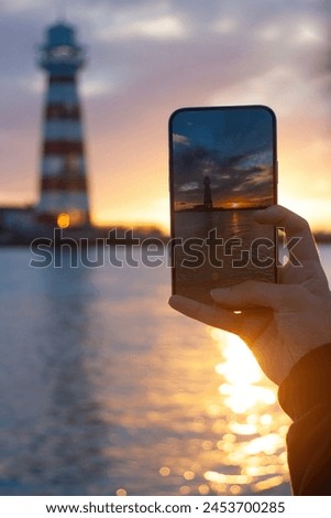 Hand holding a mobile phone and taking a picture of the lighthouse at sunset on a tropical island.