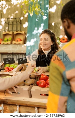 Smiling woman vendor offering customer to try out small piece of organic apple while selling fresh natural fruits and vegetables at harvest fair festival, selective focus. Tasting during shopping. Royalty-Free Stock Photo #2453728963
