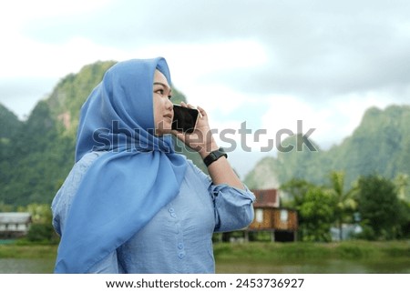 An Asian model in a hijab wearing a blue shirt is posing in nature with a backdrop of mountains and water in the countryside while holding a mobile phone