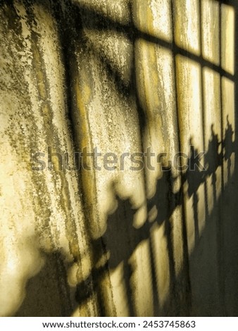 The shadow of a fence railing on a rough yellow wall, creating a visually intriguing pattern. The play of light and shadow adds depth and texture to the simple wall surface, adding an artistic element