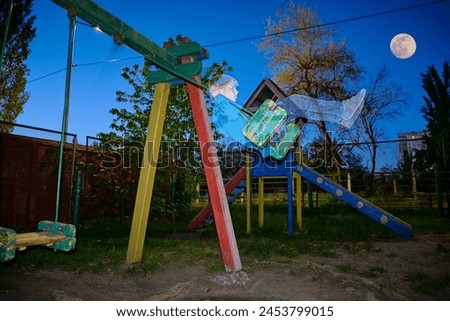 Abandoned playground with a transparent boy swinging on a swing set in the moonlight. Royalty-Free Stock Photo #2453799015