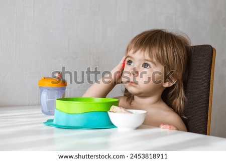 A young toddler girl with curly hair, dressed in pink overalls, sits at a wooden table. She looks thoughtfully at a plate, cup, and spoon set before her. Ideal for stock photo use.