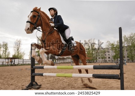 Horse and rider over jump, dynamic equestrian event. Practice session. Female jockey in uniform. Equestrian sport. Horseback riding school Royalty-Free Stock Photo #2453825429