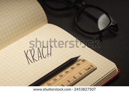 A handwritten inscription "Krach" on a grille of an open notebook on a wooden countertop, next to a black pencil, lighting of light. (selective focus), translation: crash