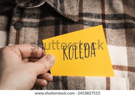 Yellow card with a handwritten inscription "Kolęda", a shaft in the hand, protruding from a brown plaid shirt (selective focus), translation: carol