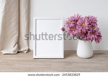 Empty white photo frame or poster mockup, vase with pink flowers bouquet on light beige wooden table, linen textile curtain and white wall background, Aesthetic still life in Nordic style.