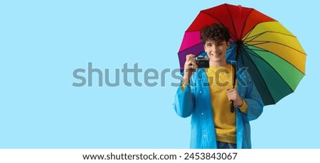 Young man in raincoat holding rainbow umbrella and photo camera on light blue background with space for text
