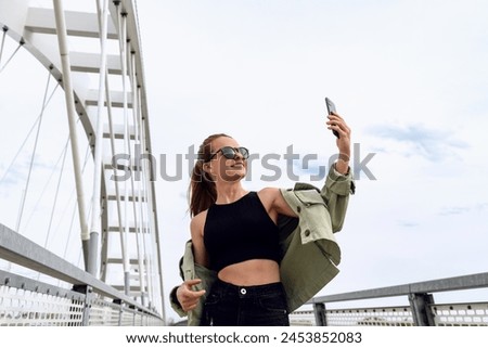 Young urban woman takes a selfie in front of city bridge.