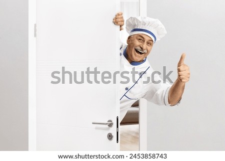 Mature chef in a uniform gesturing thumbs up behind a door and smiling