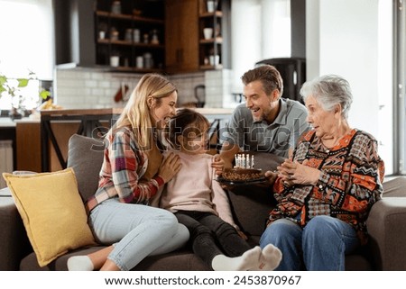 Heartwarming scene unfolds as a multi-generational family gathers on a couch to present a birthday cake to a delighted grandmother, creating memories to cherish Royalty-Free Stock Photo #2453870967