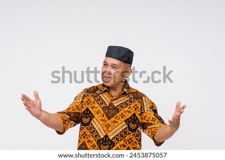 Exuberant Indonesian man in traditional batik shirt and kopiah hat elaborating, describing something large. Isolated on white background. Royalty-Free Stock Photo #2453875057