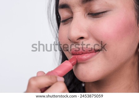 Close-up image of a Southeast Asian woman applying pink lipstick, isolated on a white background. She touches up her makeup with a smile.