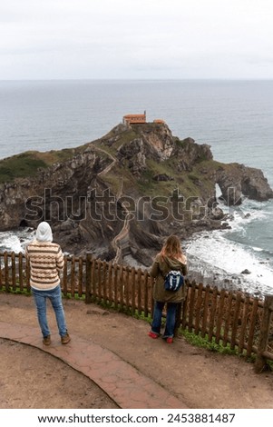 Vertical shot of a couple with their backs to San Juan de Gaztelugatxe, a tourist monument on the Basque coast, taking pictures of the incredible landscape.