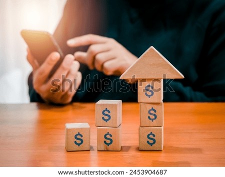 Big arrowhead wooden on increase cube blocks, business growth graph with dollar money icon while businessman holding smartphone. Investment online, stock trader, digital marketing work passive income.