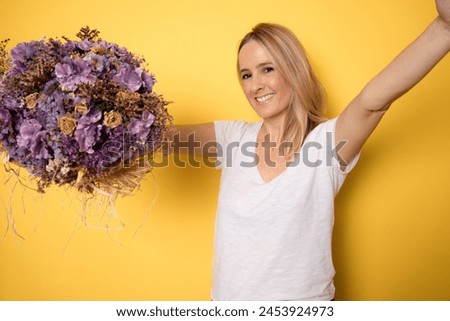 Young smiling woman holding flowers while stretching out hands to give hug with cute loving smile isolated on yellow background.
