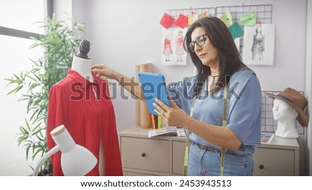 A mature hispanic woman tailor with tablet engages in her craft at a well-organized atelier, surrounded by design sketches and a mannequin dressed in red.