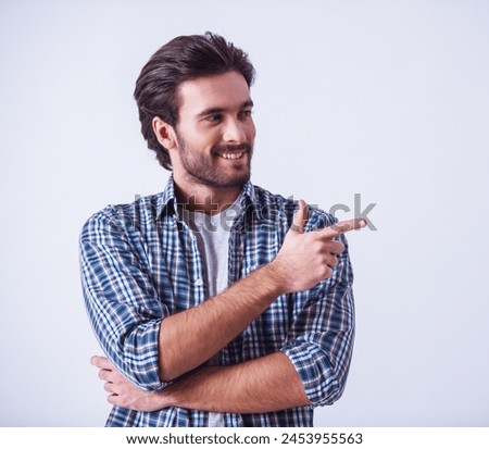 Handsome bearded man in casual clothes is pointing away and smiling, on light background