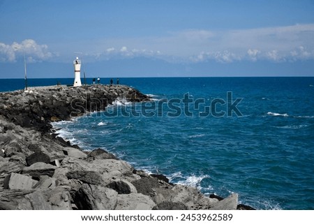 Blue partly cloudy sky on the left side black rocks white lighthouse and blue sea at the end of the rocks Royalty-Free Stock Photo #2453962589