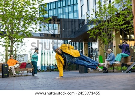 Young Stylish B-boy Breakdancing On Street Among Modern Buildings In Urban Area. Fashionable Group Of Friends Chilling On Background, Supporting Breakdance Performer Practising Choreography. Royalty-Free Stock Photo #2453971455