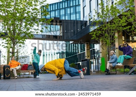 Young Stylish B-boy Breakdancing On City Street Among Buildings In Urban Area. Fashionable Group Of Friends Chilling On Background, Supporting Breakdance Performer Practising Choreography. Royalty-Free Stock Photo #2453971459