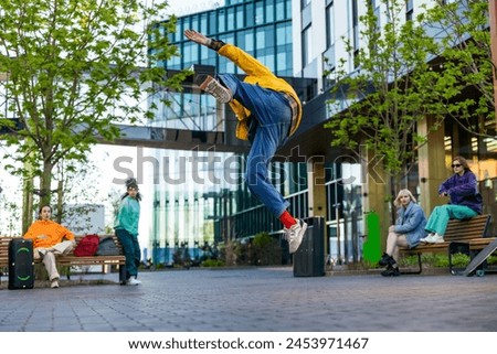 Young Stylish B-boy Breakdancing On City Street Among Modern Buildings In Urban Area. Fashionable Group Of Friends Chilling On Background, Supporting Breakdance Performer Practising Choreography. Royalty-Free Stock Photo #2453971467