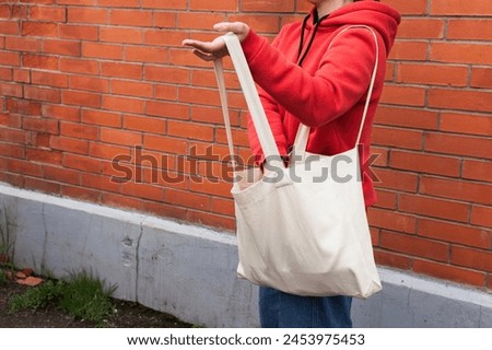 Woman in red hoodie holding tote canvas blank eco bag on street brick wall background. Female consumer hold white textile shopper. template or place for your design, logo, text