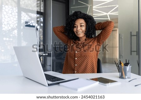 A young African American woman in a polka dot shirt stretches comfortably at her modern office desk, exuding a relaxed, confident vibe. Royalty-Free Stock Photo #2454021163