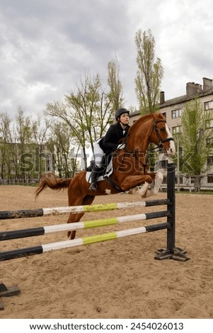 Focused rider clearing jump with horse, vertical shot. Female jockey in uniform. Equestrian sport. Horseback riding school Royalty-Free Stock Photo #2454026013