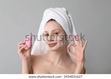 Young woman washing face with brush and cleansing foam on grey background