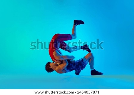 Young men, wrestlers compete in a high-energy match, demonstrating skill and strategy against blue background in neon light. Concept of combat sport, martial arts, competition, tournament, athleticism