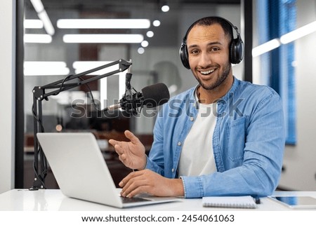 Portrait of a smiling young Muslim man sitting in headphones in the office at the table, talking online through a microphone, looking at the camera.